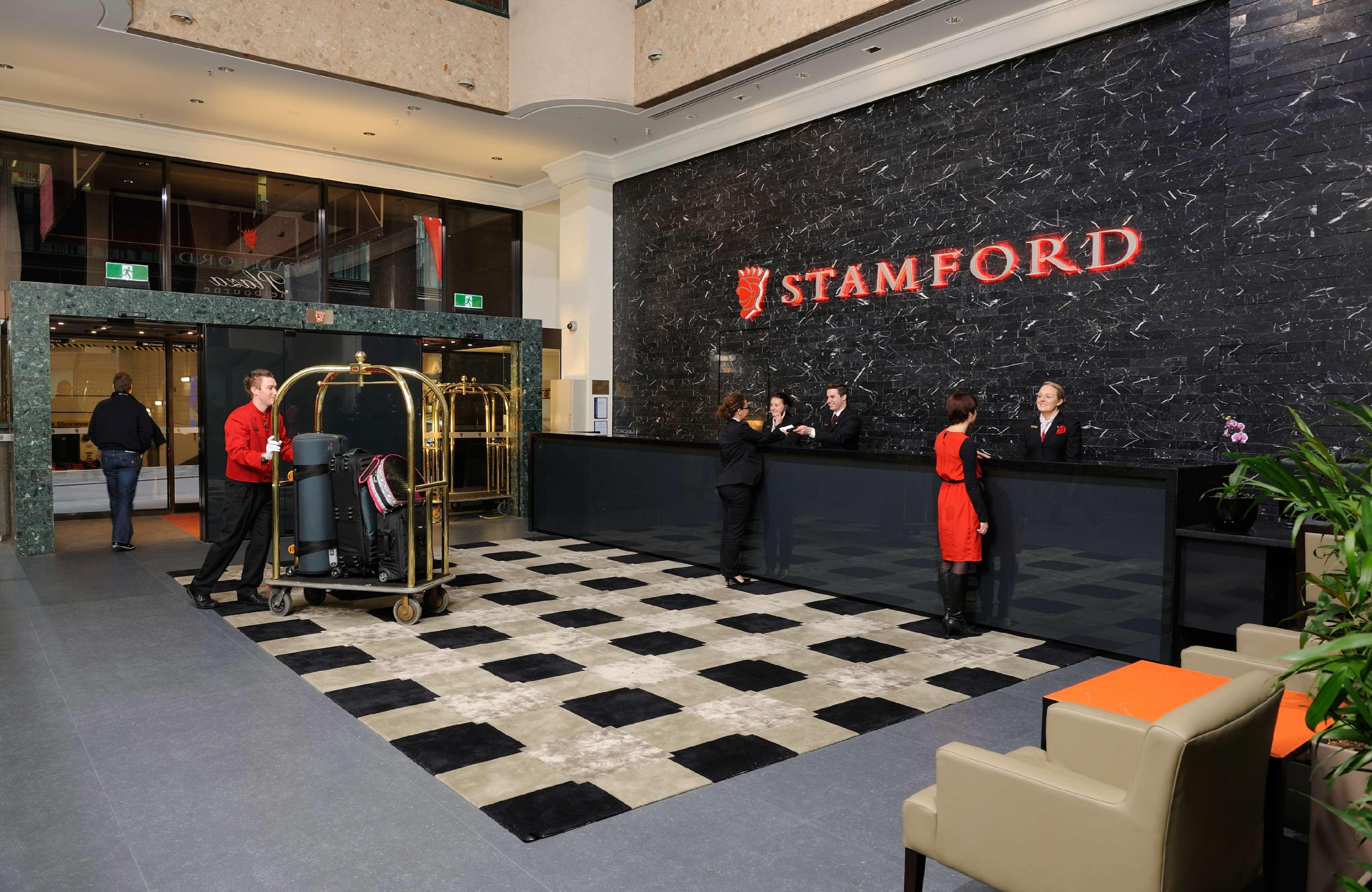Hotel Stamford Plaza Ville de Melbourne Intérieur photo The photo shows the interior of a hotel lobby, specifically the Stamford Hotel. There is a large reception desk where several staff members are attending to guests. In the foreground, a bell cart loaded with luggage is positioned beside the reception