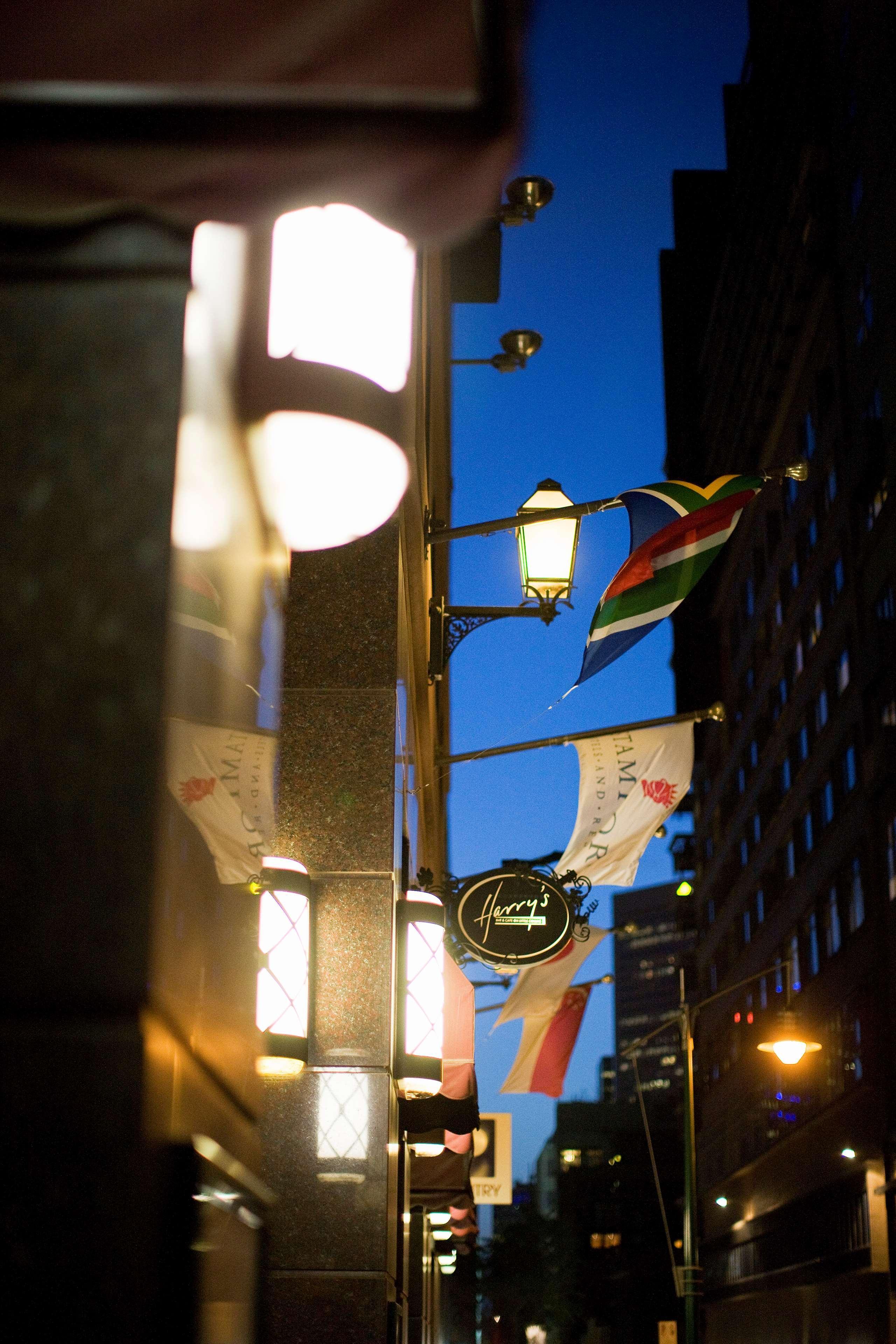 Hotel Stamford Plaza Ville de Melbourne Extérieur photo The photo features a street view of a building at dusk. Prominently displayed are several flags, including a South African flag, which are fluttering in the wind. There is a sign that reads "Harry's," likely indicating the name of a business or estab