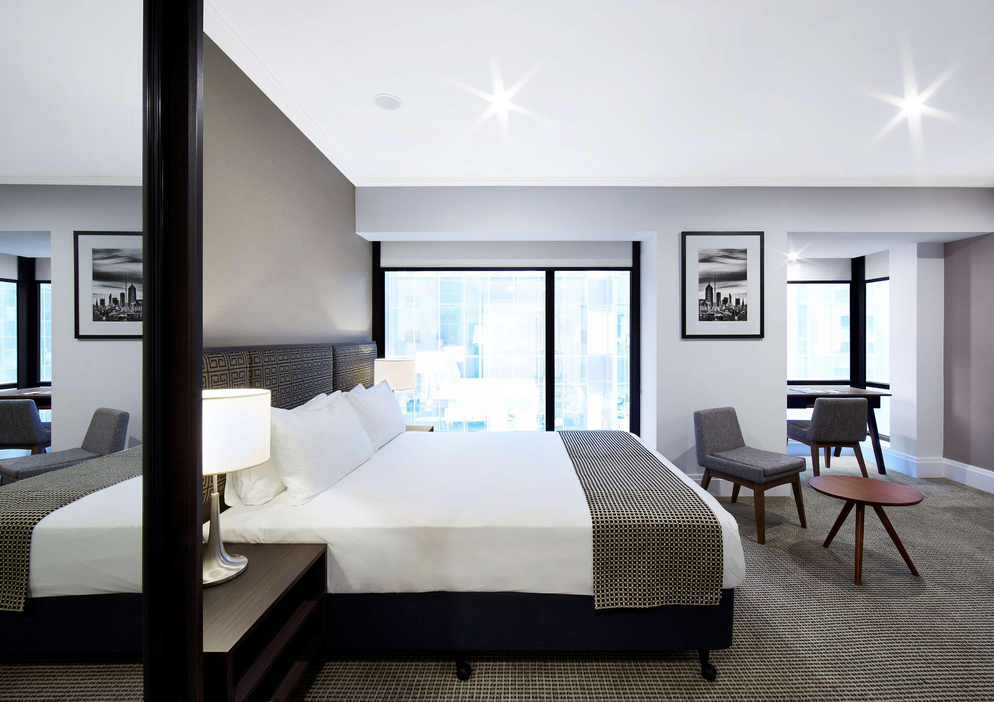 Hotel Stamford Plaza Ville de Melbourne Extérieur photo The image shows a modern hotel room featuring a large bed with white linens and a gray throw blanket. There are two windows that allow natural light to enter the space, and the walls are painted in a light color. In addition to the bed, there are two
