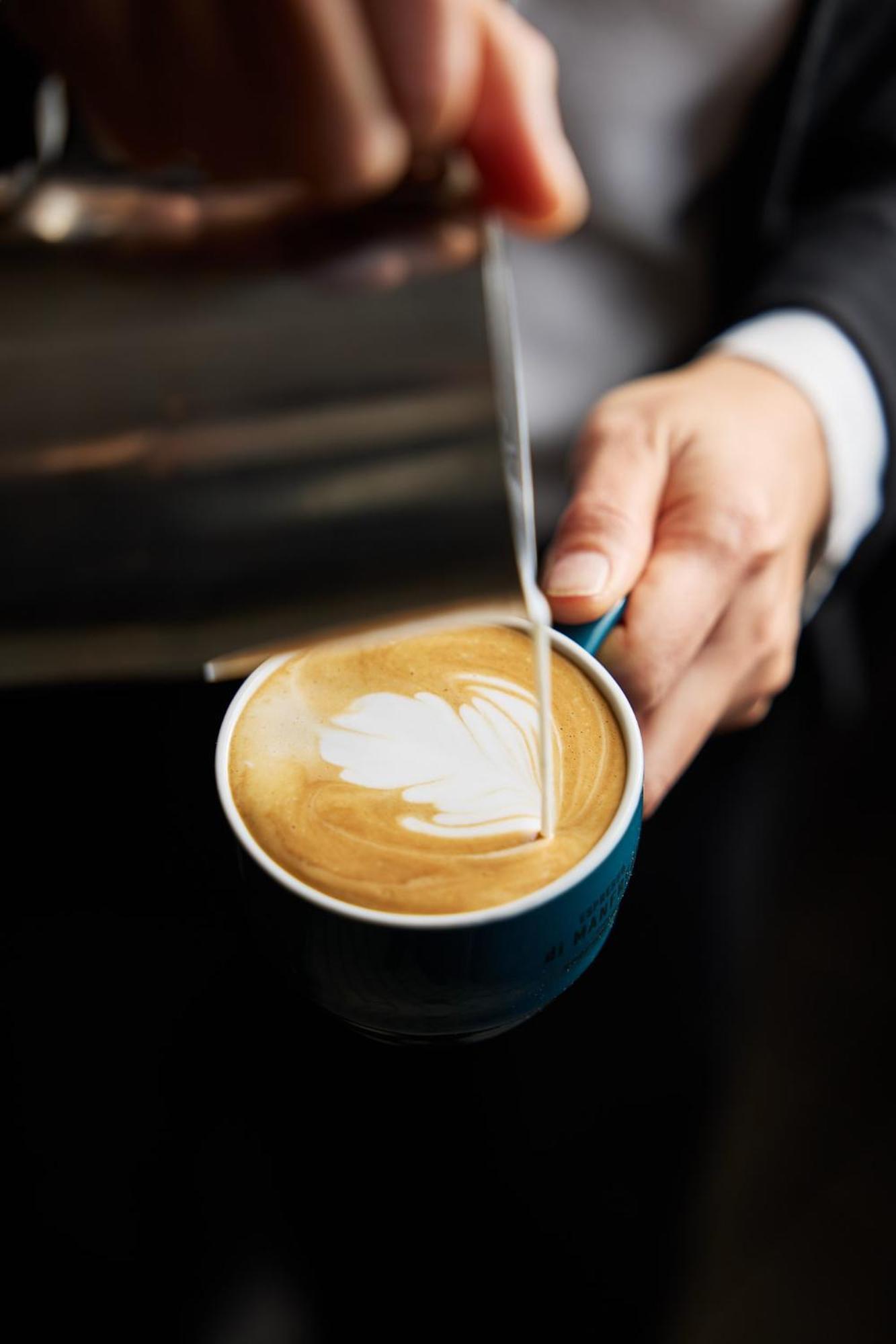 Hotel Stamford Plaza Ville de Melbourne Extérieur photo The photo shows a close-up of a hand holding a cup of coffee with a latte art design on the surface. The coffee appears to be a creamy, caramel color, likely a latte or cappuccino. A small stream of milk is being poured into the cup, creating a heart