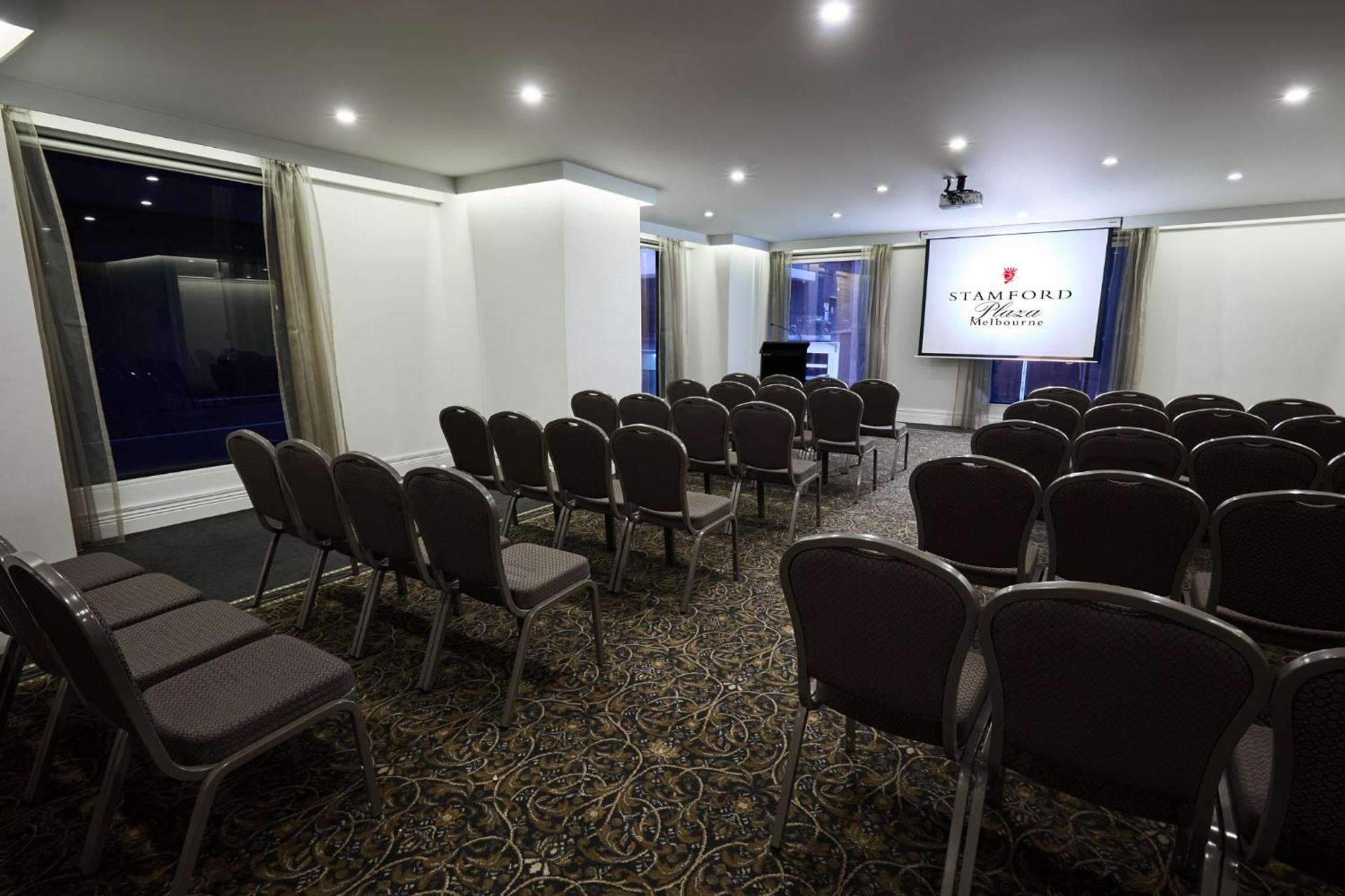 Hotel Stamford Plaza Ville de Melbourne Extérieur photo The image shows a conference or meeting room set up for an event. There are rows of gray chairs arranged facing a large screen displaying the name "Warford House." The room is well-lit with overhead lighting and has large windows covered with light-c