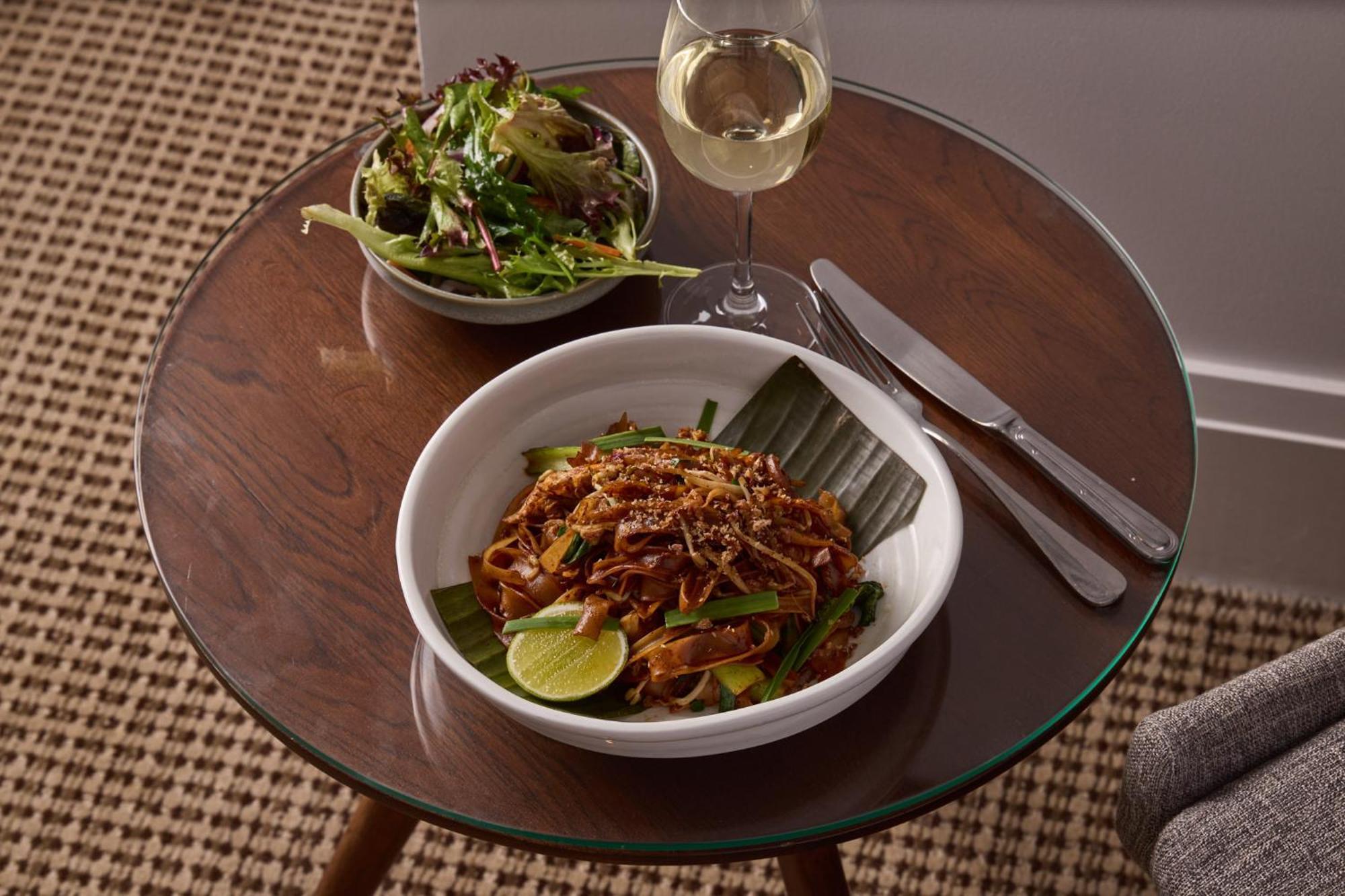 Hotel Stamford Plaza Ville de Melbourne Extérieur photo The photo shows a meal served on a wooden table. In the foreground, there is a bowl of stir-fried noodles garnished with green onions and a lime wedge, set atop a banana leaf. Next to it, there is a bowl of mixed green salad. A glass of white wine si