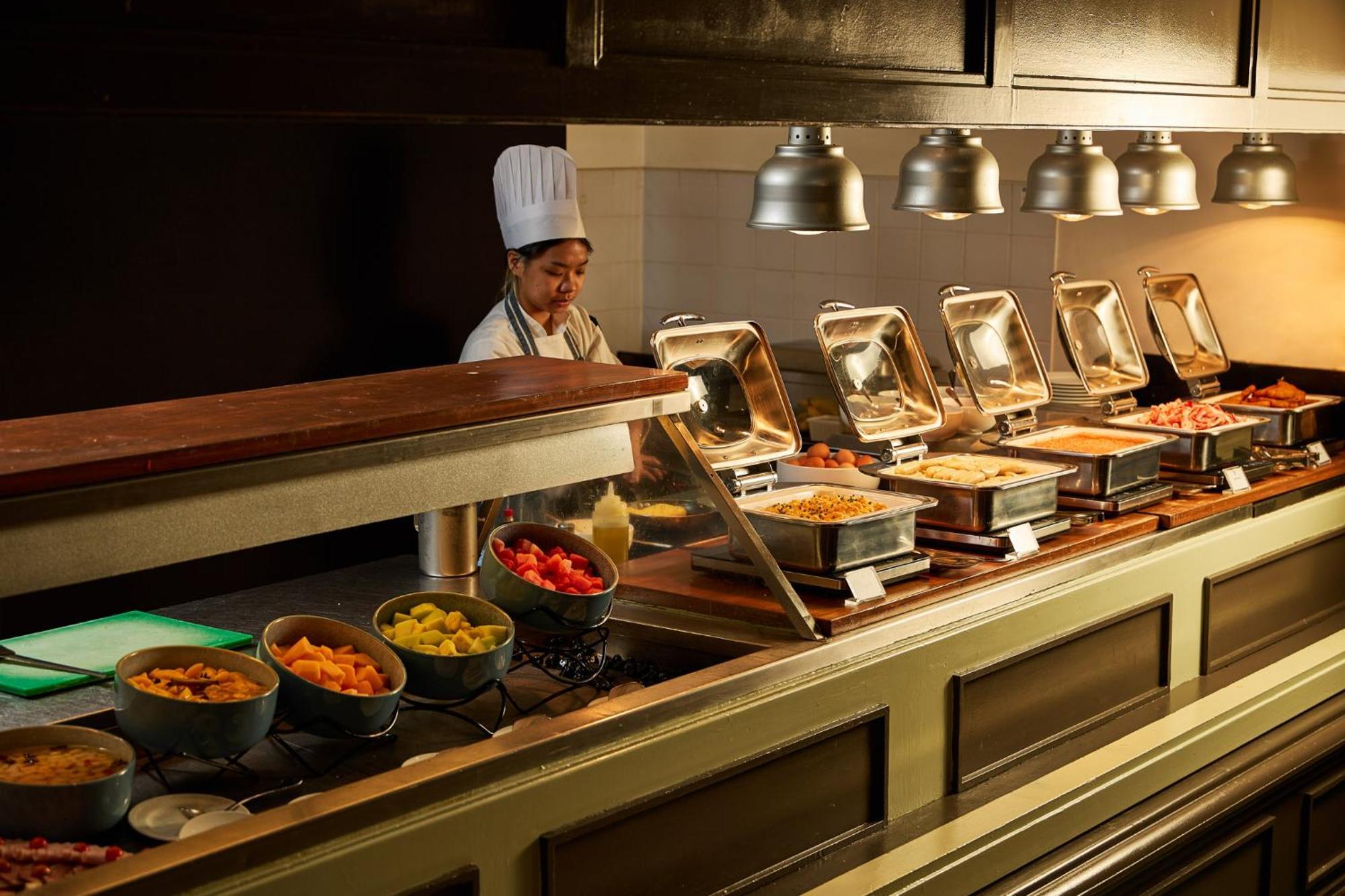 Hotel Stamford Plaza Ville de Melbourne Extérieur photo The photo shows a buffet setup in a dining area. There is a chef in a white uniform and hat, standing behind a counter filled with various food options. The buffet features several metal serving trays containing different dishes, with warming lights 
